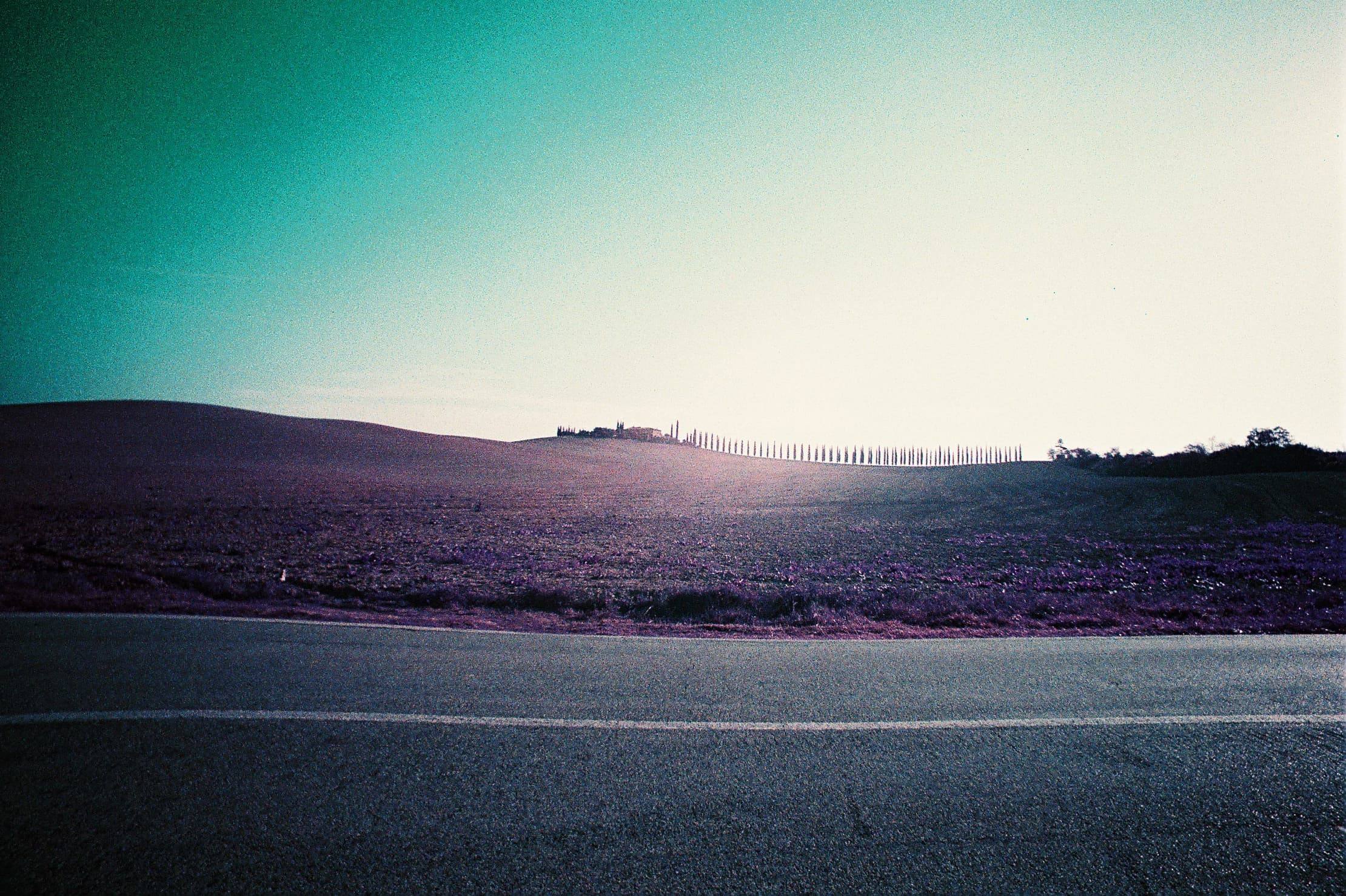 Purple-toned film photograph of a road in Tuscany: a low ploughed hill, a bright sky and a distant line of cypress trees.