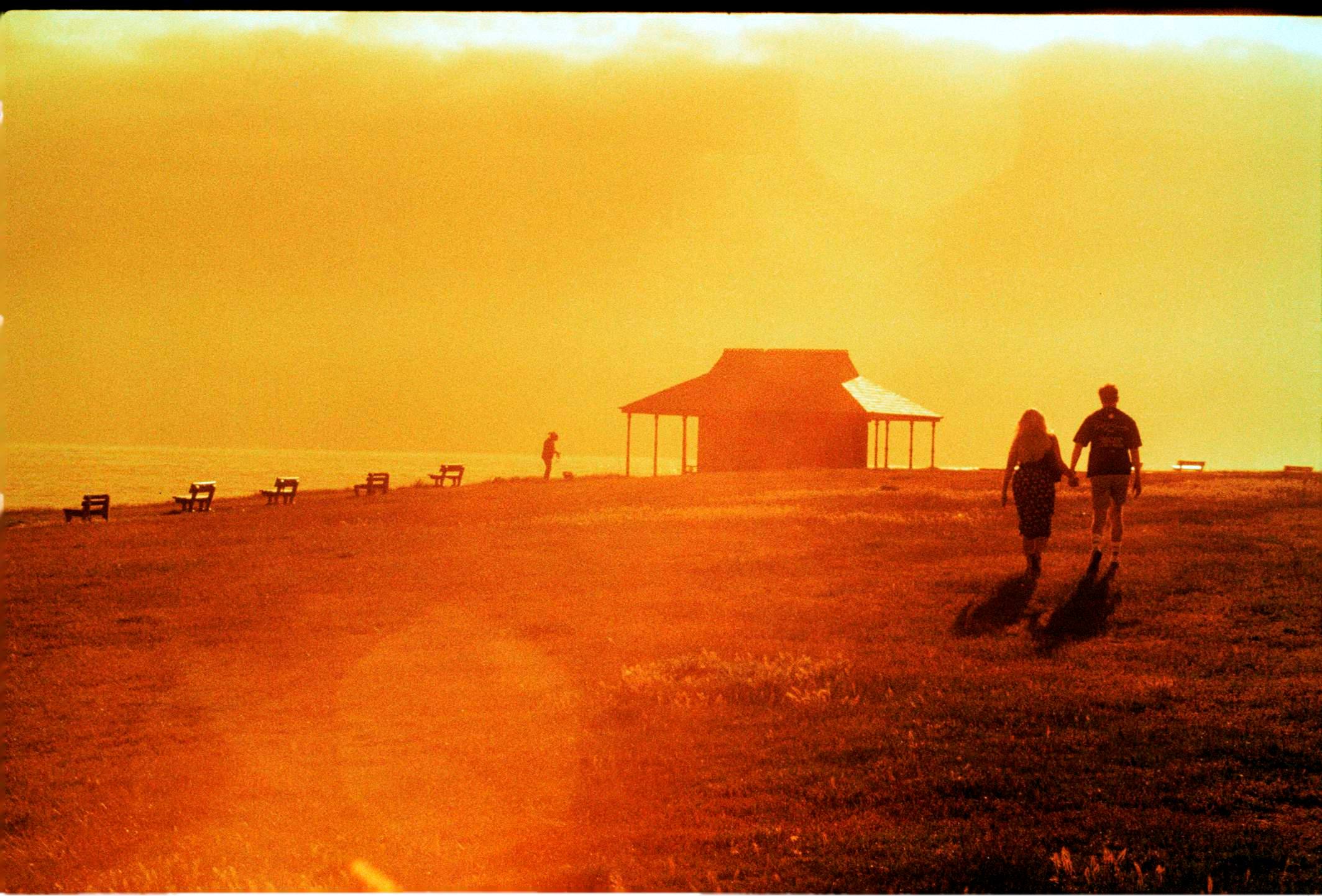 Rescale film photograph washed in orange and yellow light leaks: a couple walking across a lawn towards a small pavilion, with the sea behind.