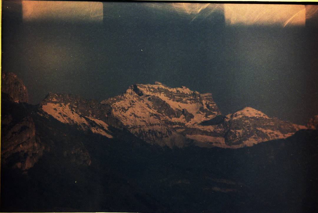 Snow-capped mountain range on expired colour film, the frame fogged with warm light leaks along its edges.