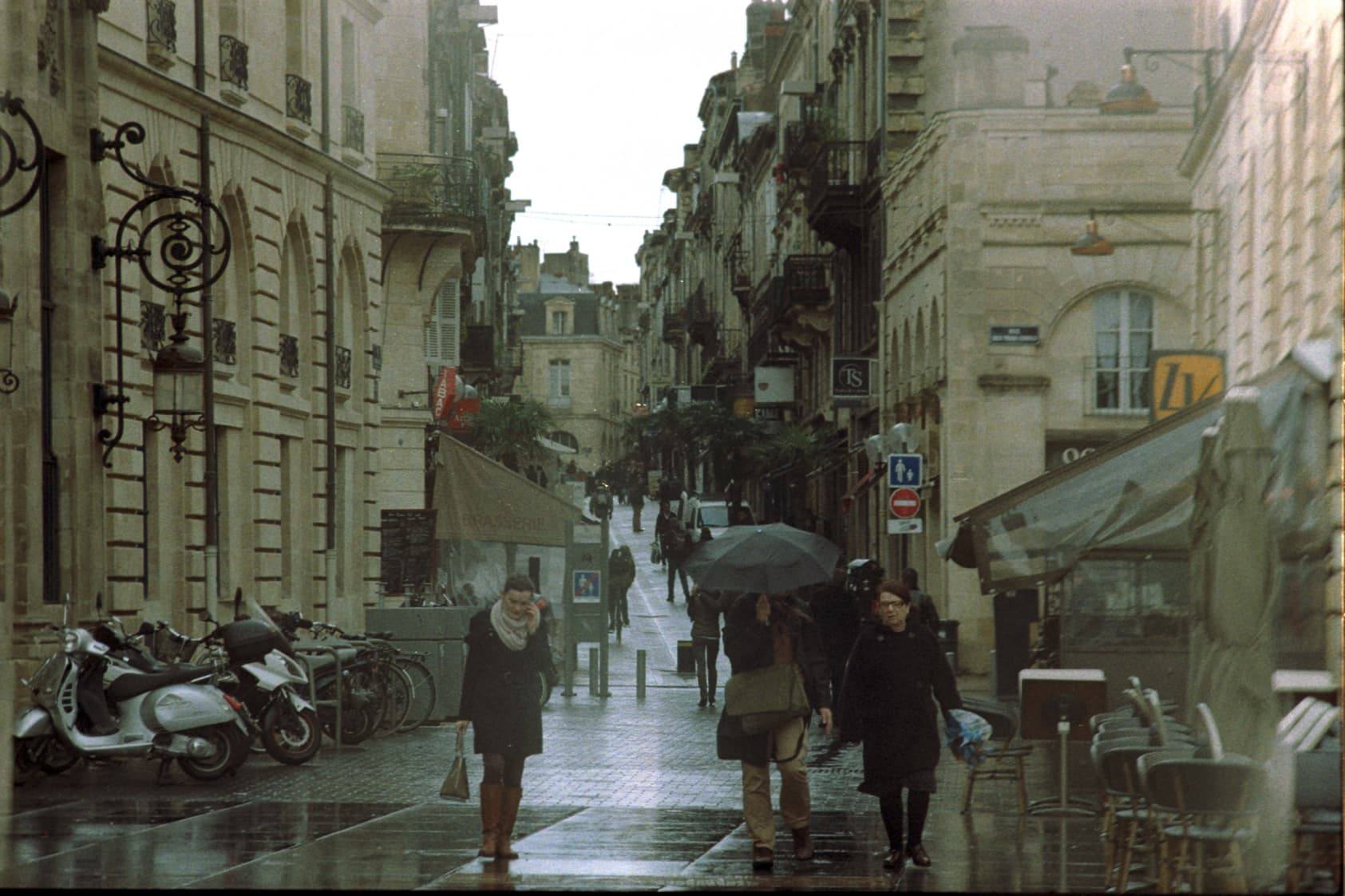 Colour film photograph of a rainy street in Bordeaux: people with umbrellas, parked scooters and tall stone buildings.
