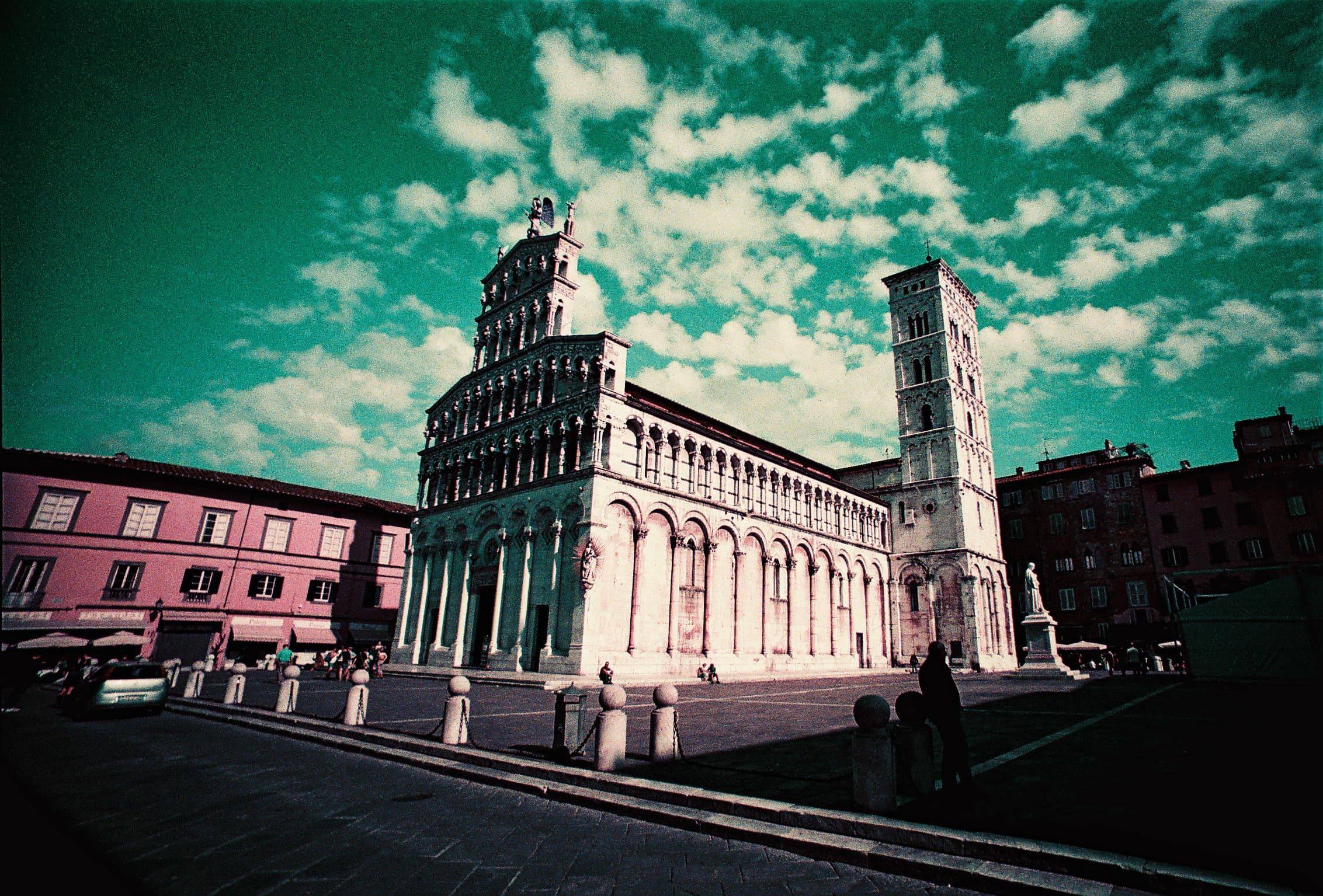 Lomochrome Purple film photograph of the church of San Michele in Lucca, its white marble façade under a teal sky with streaked clouds.