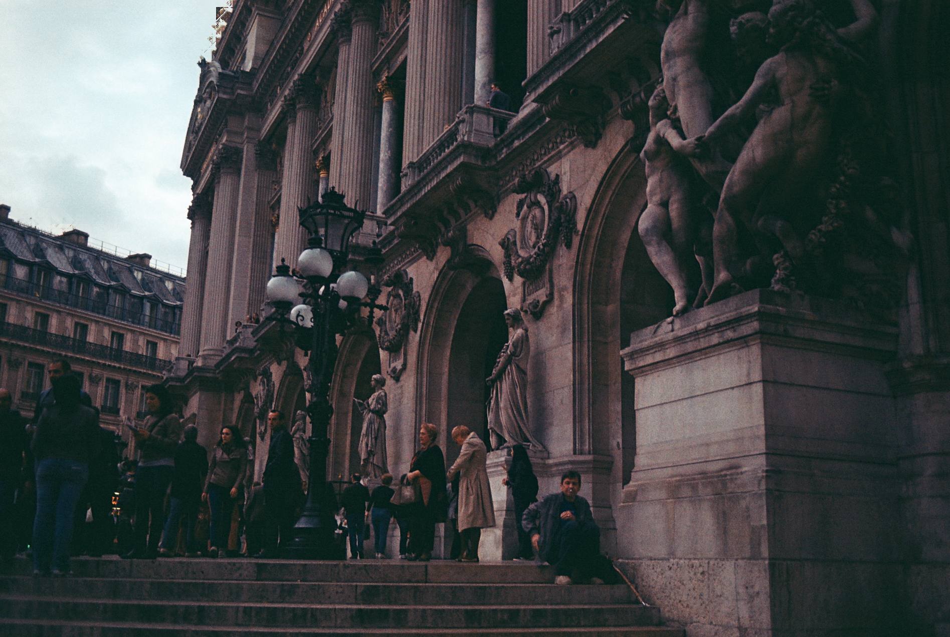 Colour film photograph of people gathered on the steps of an ornate Paris building, with a stone statue and a lamp post at the entrance.