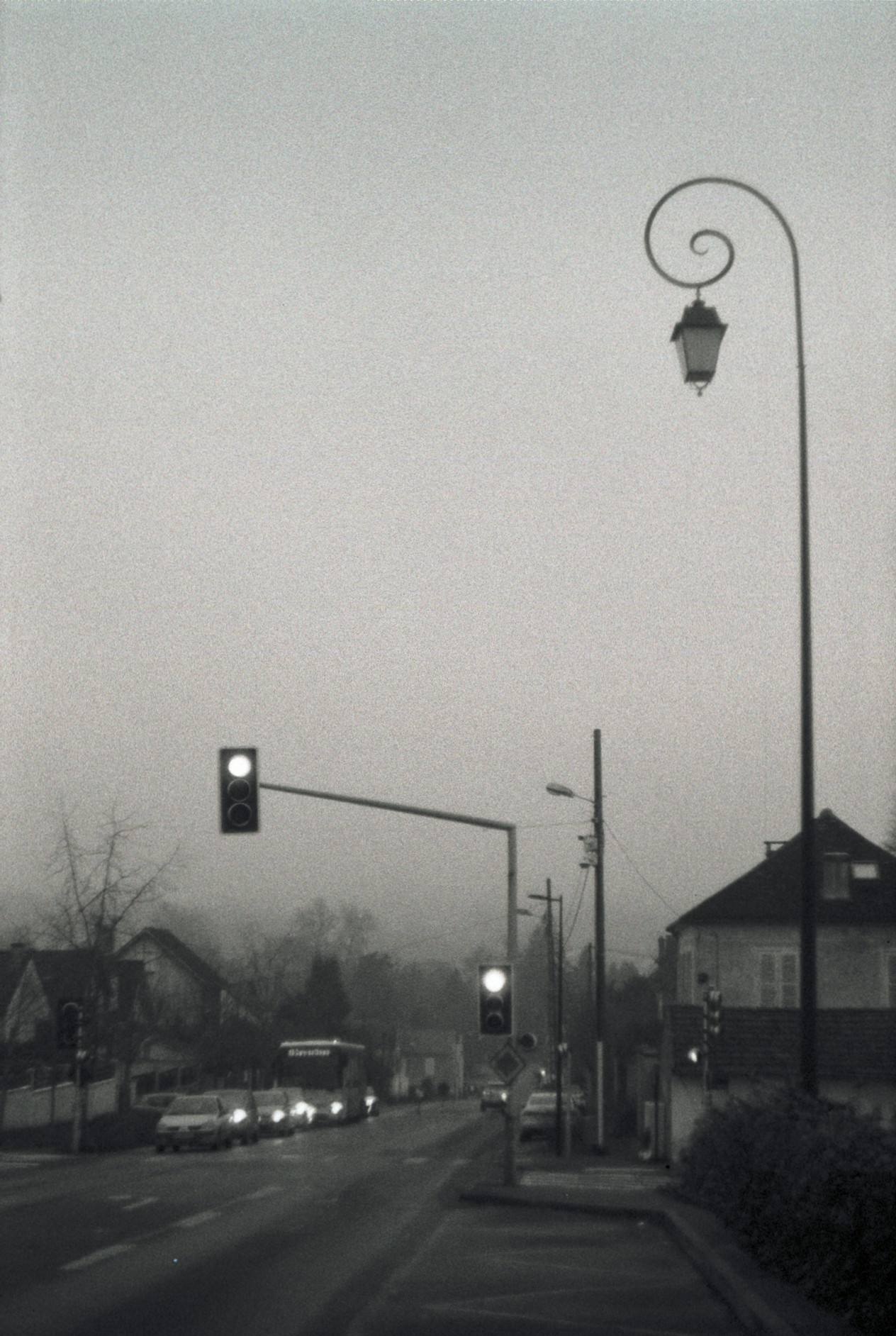 Black-and-white film photograph of a curved street lamp and traffic lights against a hazy dusk sky, cars and houses below.