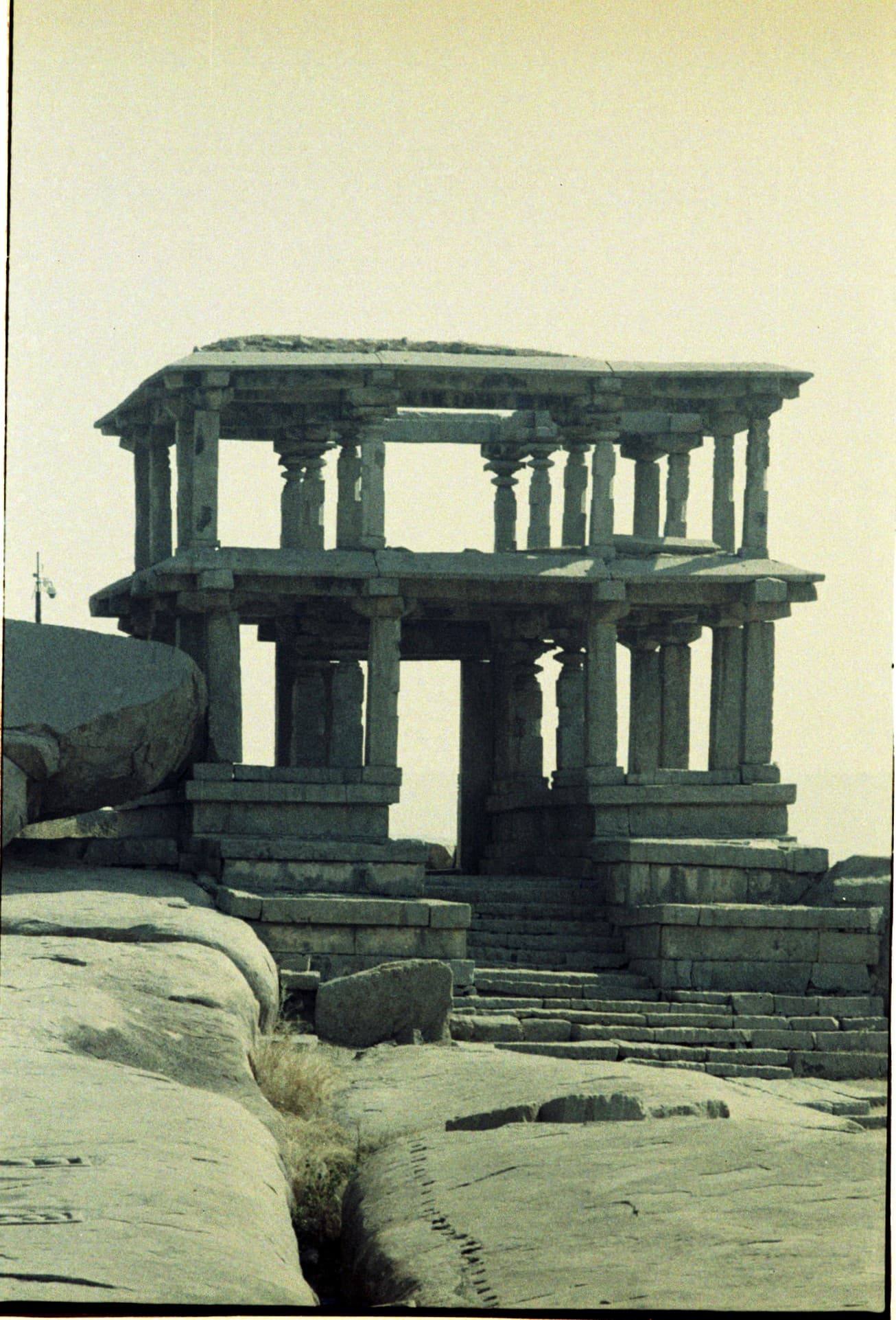 Colour film photograph of a two-storey stone pillared pavilion in Hampi standing on bare rock under a pale sky.