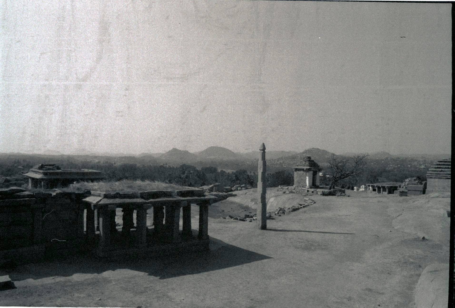 Black-and-white film photograph of the rocky landscape at Hampi: a stone pavilion, a tall pillar and boulder hills in haze.