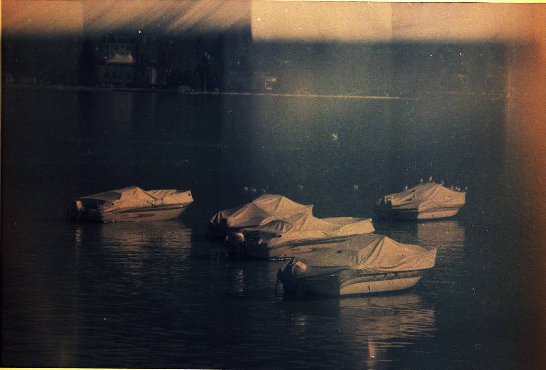 Small covered boats moored on still water at dusk on expired colour film, the frame fogged with warm light leaks.