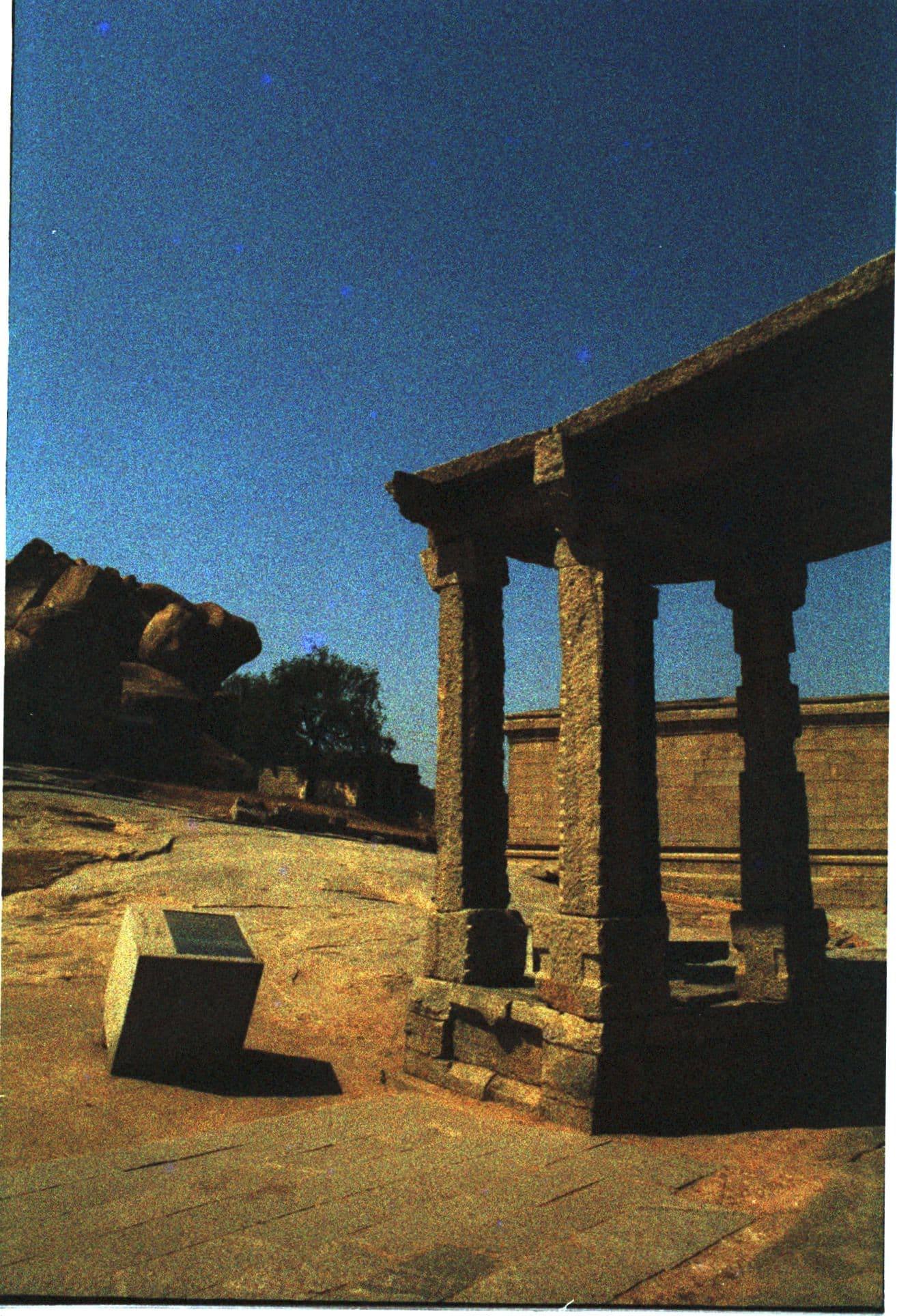 Colour film photograph of ancient stone pillars at Hampi against a deep blue sky, a carved block in the foreground.